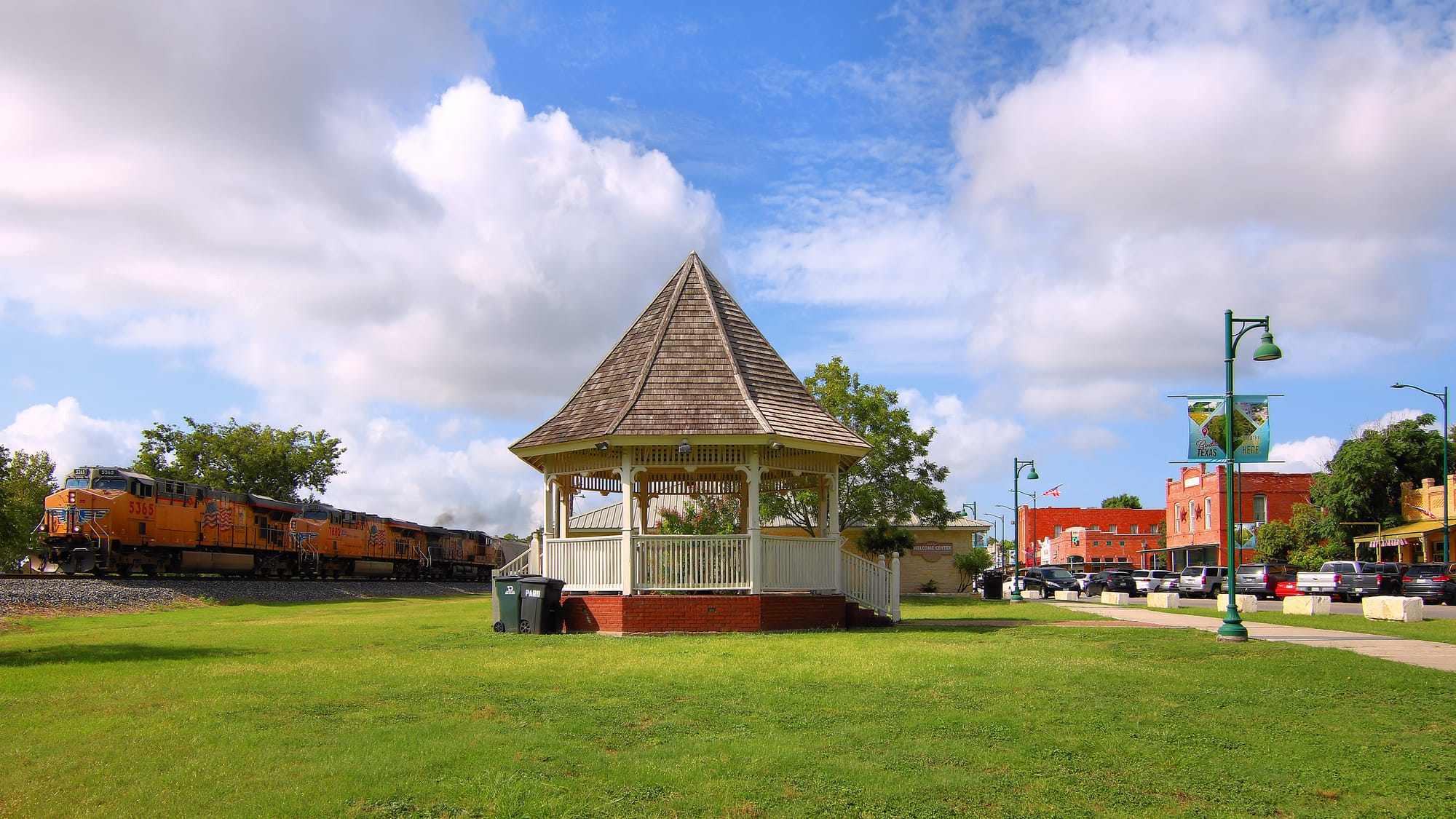 The historic downtown gazebo in Buda, Texas, with Main Street storefronts, a "Buda, Texas" welcome banner, and a passing train.